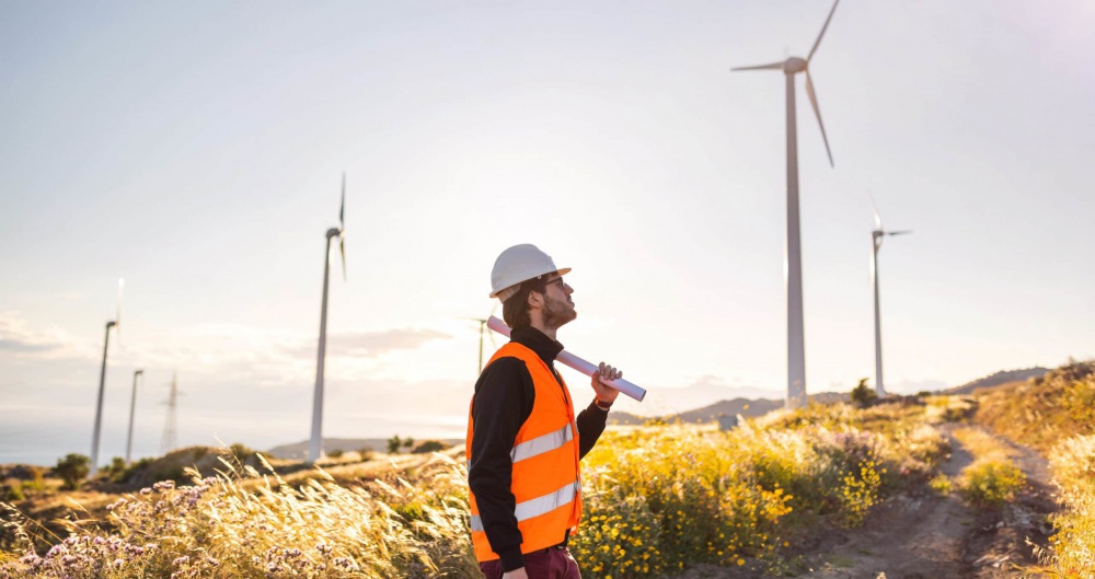 A windmill worker donning a safety vest and helmet while holding a planning document looks up at wind turbines in a field.